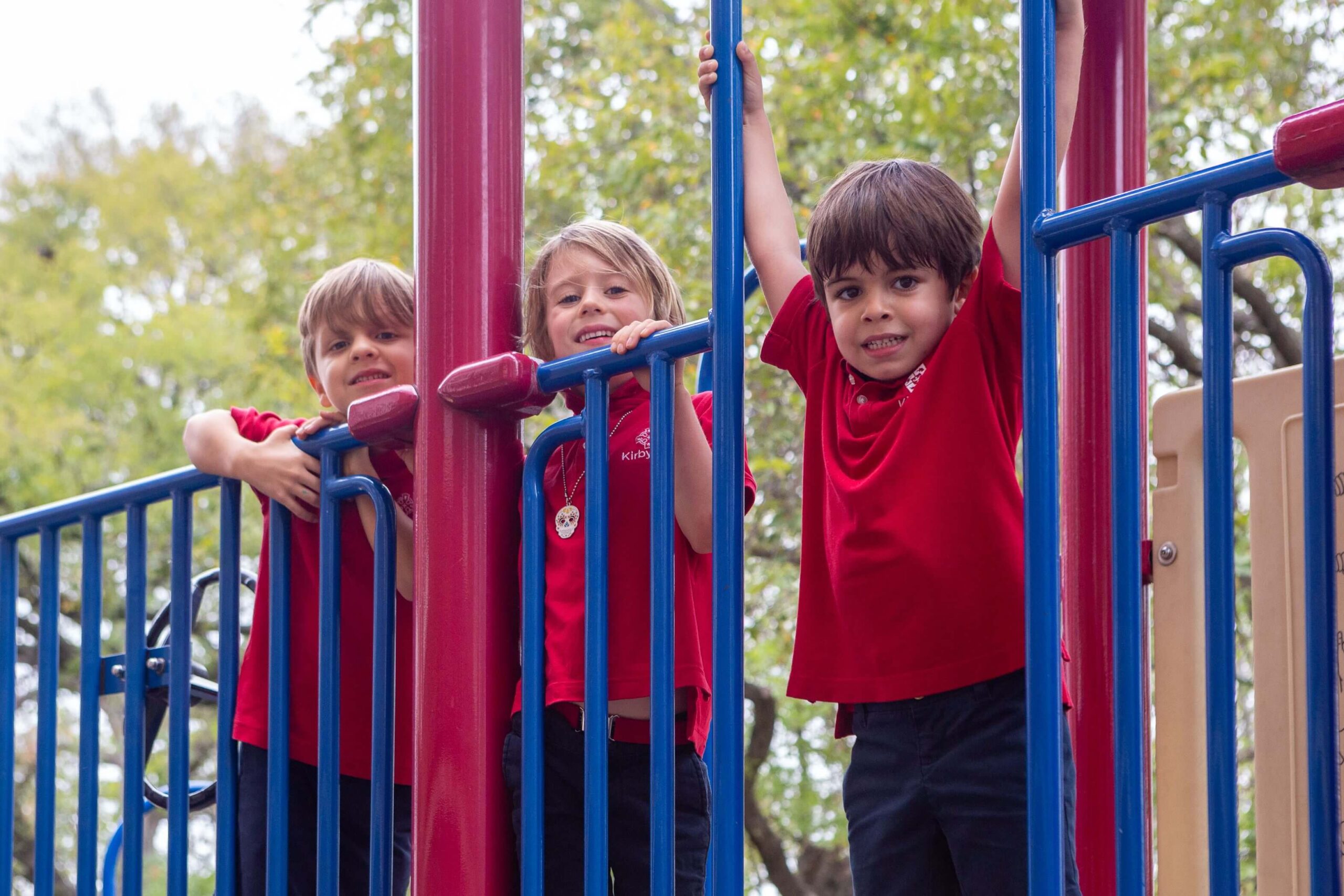 Three young children in red shirts playing on a colorful playground structure outdoors.