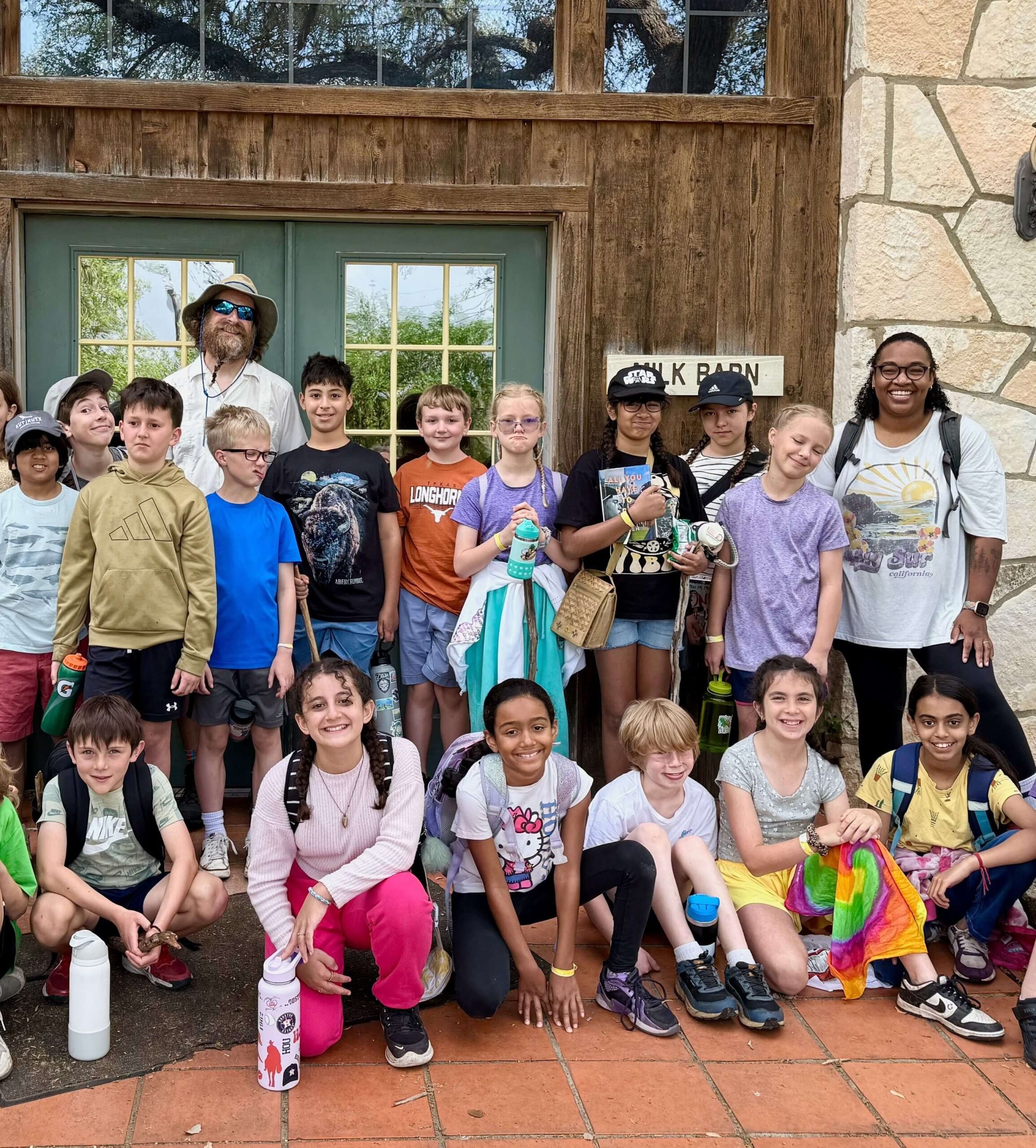 Group of children and two adults smiling outdoors, posing in front of a rustic building.