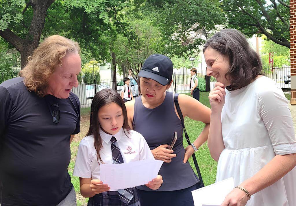 Girl in school uniform shows paper to three adults in a park setting.